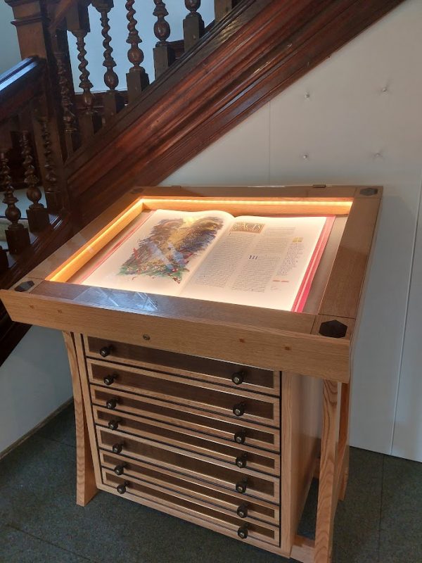 An image of the display case with the seven volumes of the St John's Bible. The exhibited edition has an illumination on the left-hand page. The display case is in front of the main, wooden staircase at Sarum College.