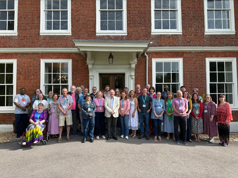 A group of smiling students and staff outside Sarum College on a sunny day